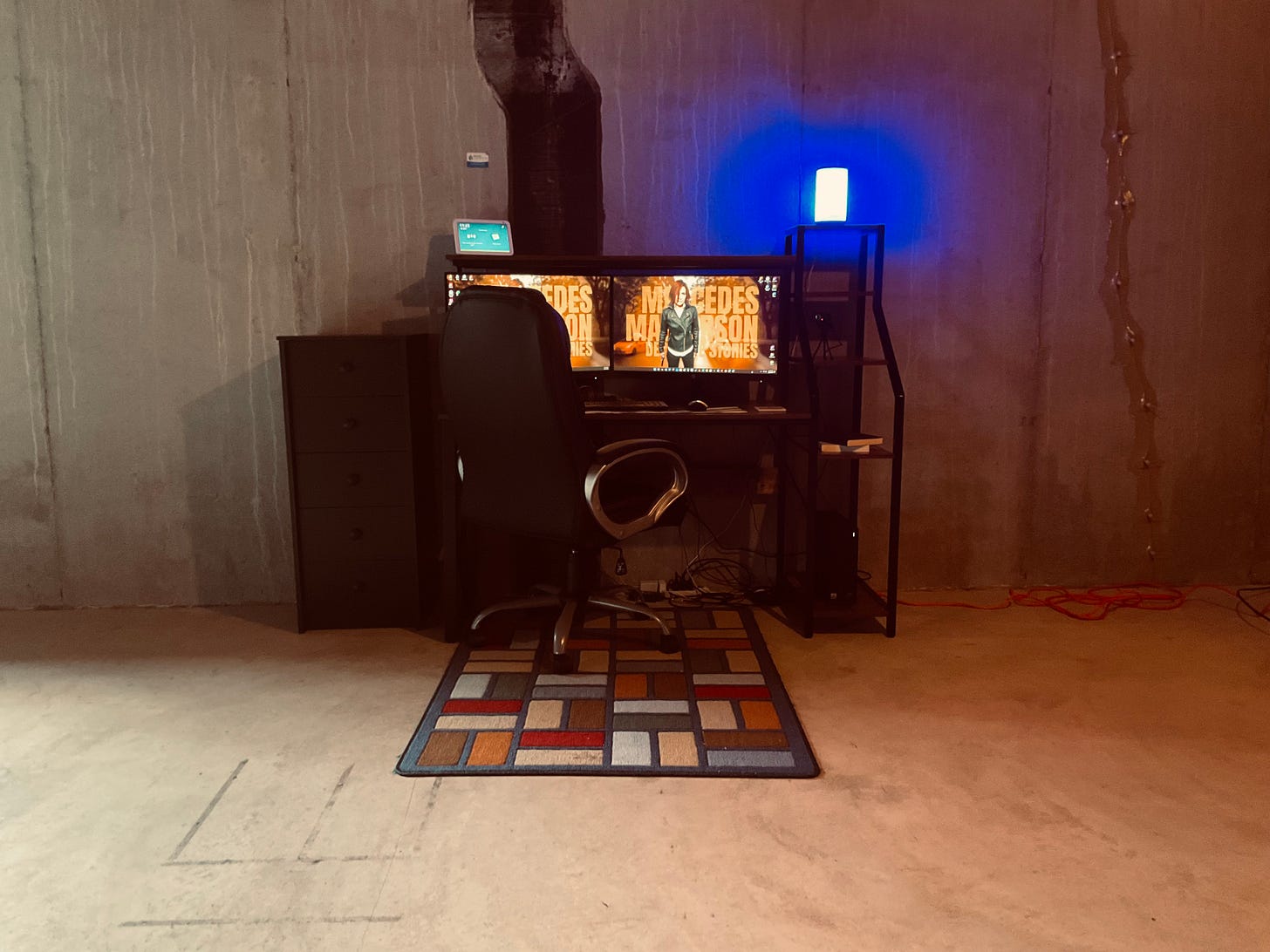 Picture of a computer desk and chair against a concrete wall in a basement. The desk has two monitors, an Echo Show 8, two books on the shelf end of the desk, a smart lamp glowing blue. There is also a tall, black cabinet to the left of the desk. Under the desk is a small rug with colorful rectangles. Picture of a computer desk and chair against a concrete wall in a basement. The desk has two monitors, an Echo Show 8, two books on the shelf end of the desk, a smart lamp glowing blue. There is also a tall, black cabinet to the left of the desk. Under the desk is a small rug with colorful rectangles.
