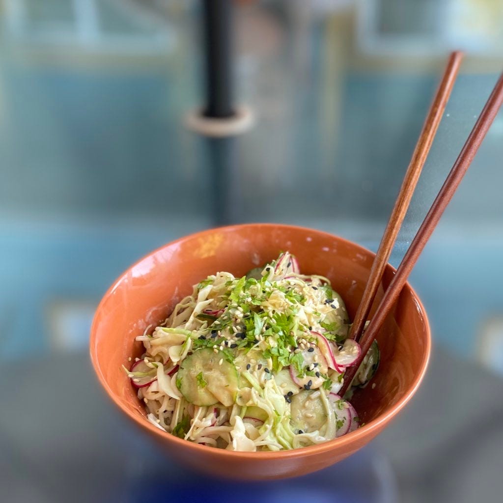 An orange bowl of ramen noodles in peanut sauce with thinly sliced radishes, cucumbers, and cabbage. On top are black and white sesame seeds and chopped cilantro. Wooden chopsticks stick out the side of the bowl.
