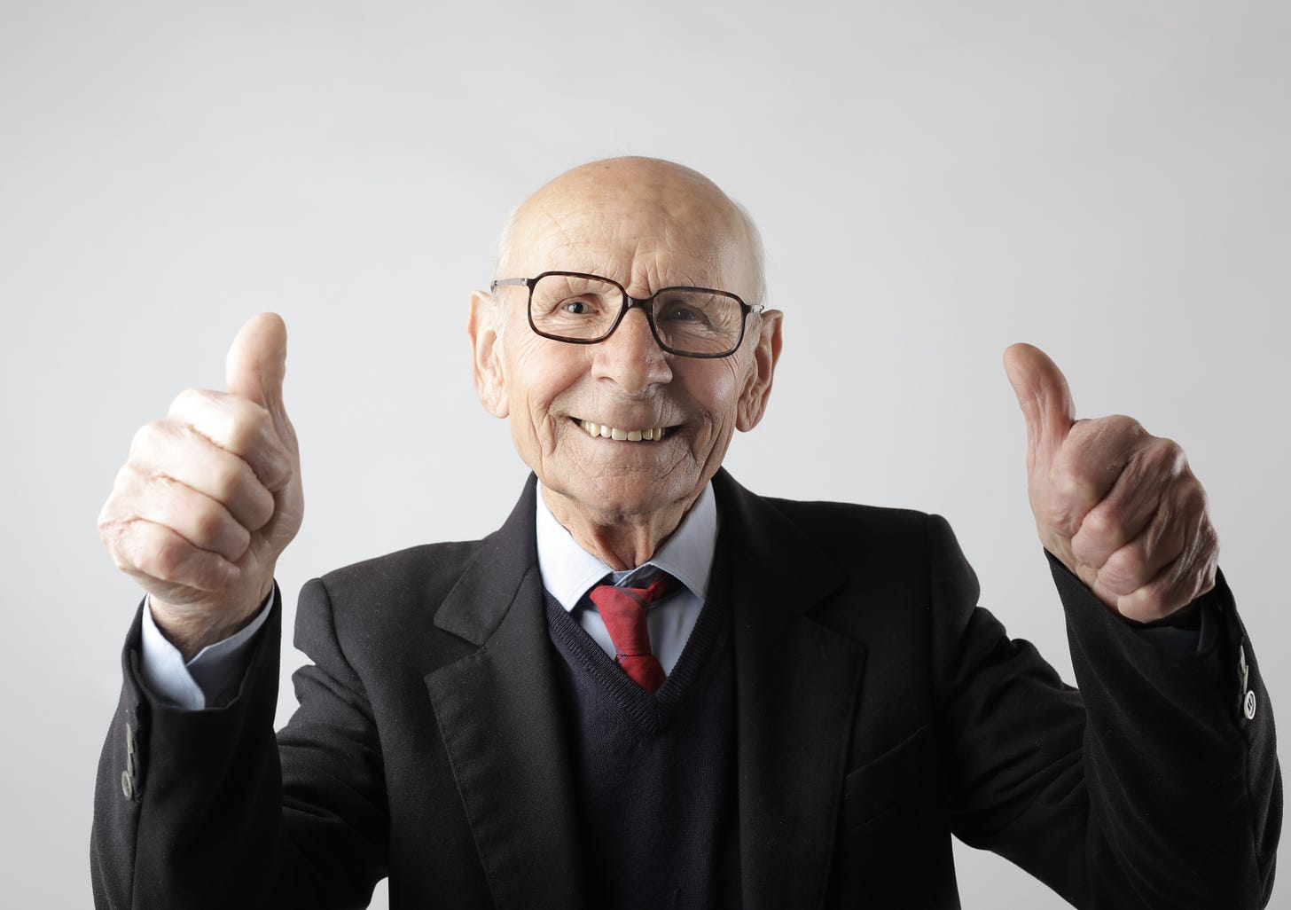 Old man in a suit smiling and giving the thumbs up with both hands on a white background