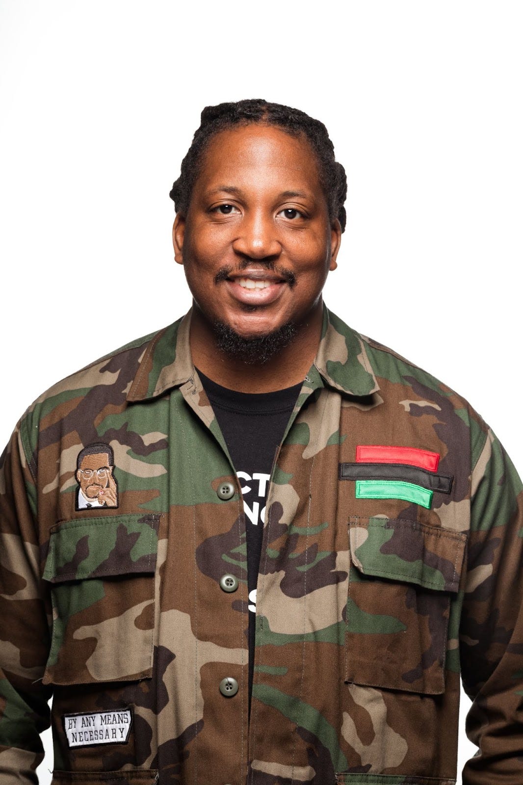 A headshot photo of Nigel Charles, smiling into the camera, wearing a camo jacket and black shirt. He has dark brown skin and his hard is braided close to his scalp. He has a mustache and on his jacket is a patch with the face of Malcolm X..