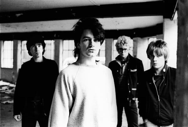 A black and white image of U2 in its early days, with Bono, center, in a light sweatshirt and the Edge, Adam Clayton and Larry Mullen Jr dressed in dark colors. 