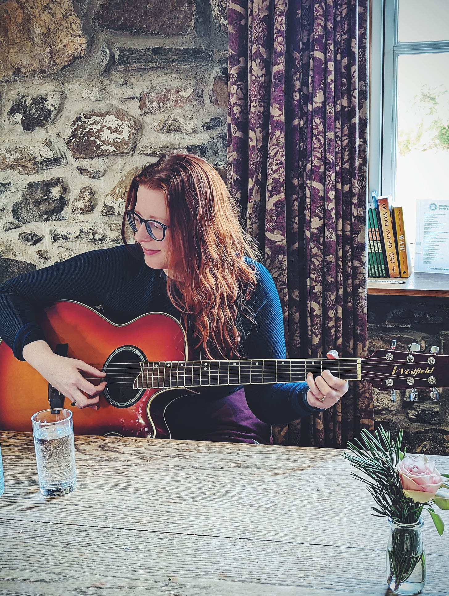 A smiling woman in her thirties is playing guitar at a long table. She is happy and content. There are books on the sill behind her and an old stone wall, and long patterened gold and plum curtains. SHe has red hair and blue glasses to match her blue top.