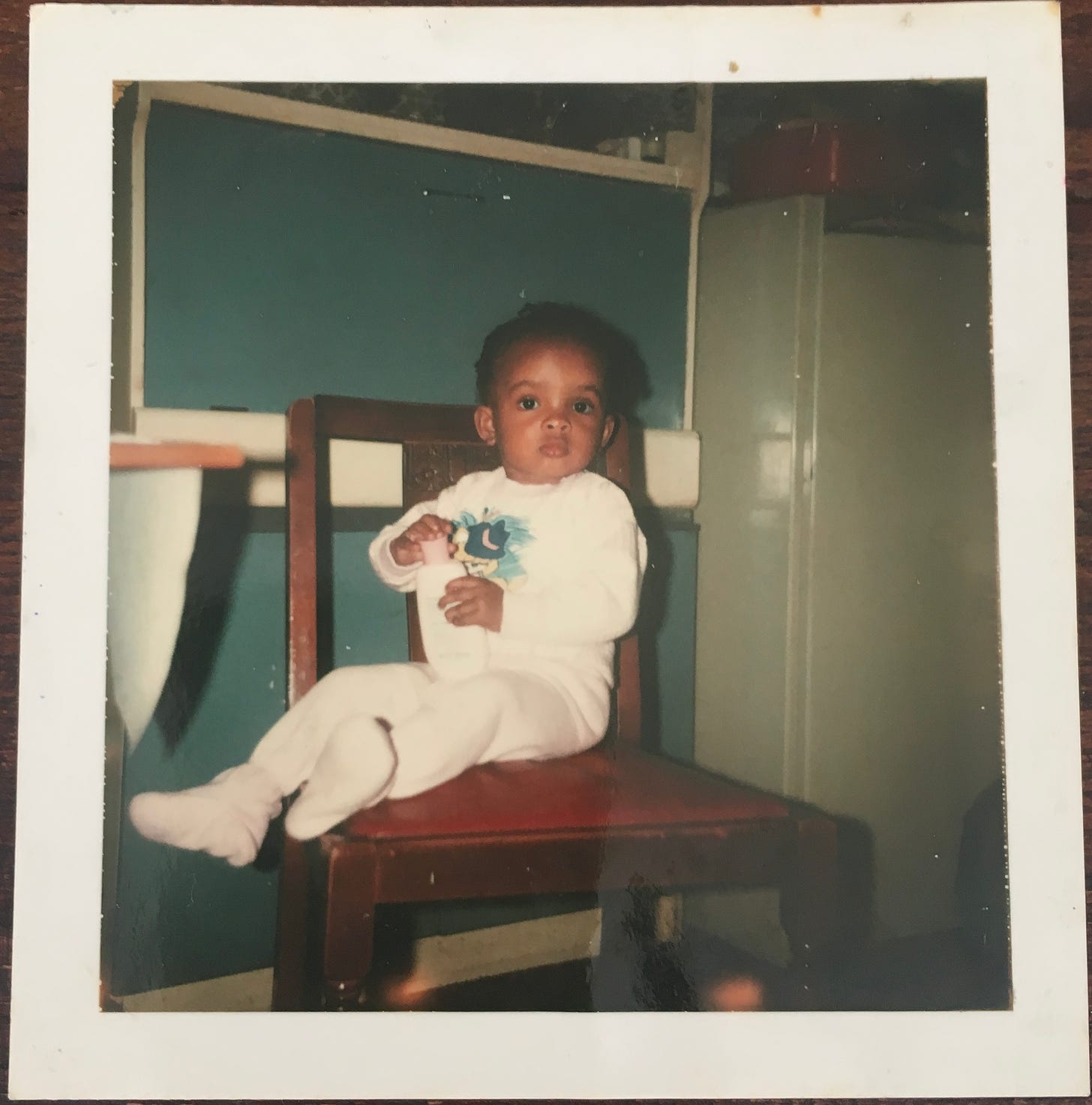 Baby Natalie Lue in a babygro sat on a dining chair holding a bottle of moisturiser