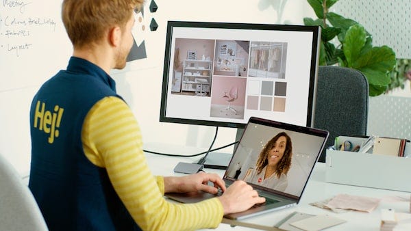 A man sits at a desk engaged in a video call with a woman via a laptop. A second screen shows images of room interiors.