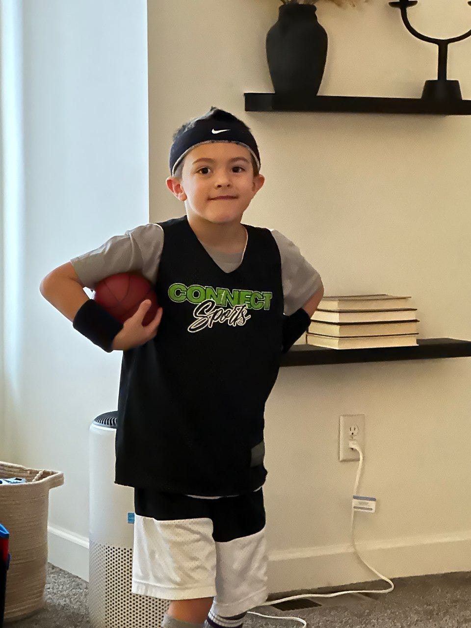 A young boy wearing basketball gear holds a basketball while standing in front of a white wall