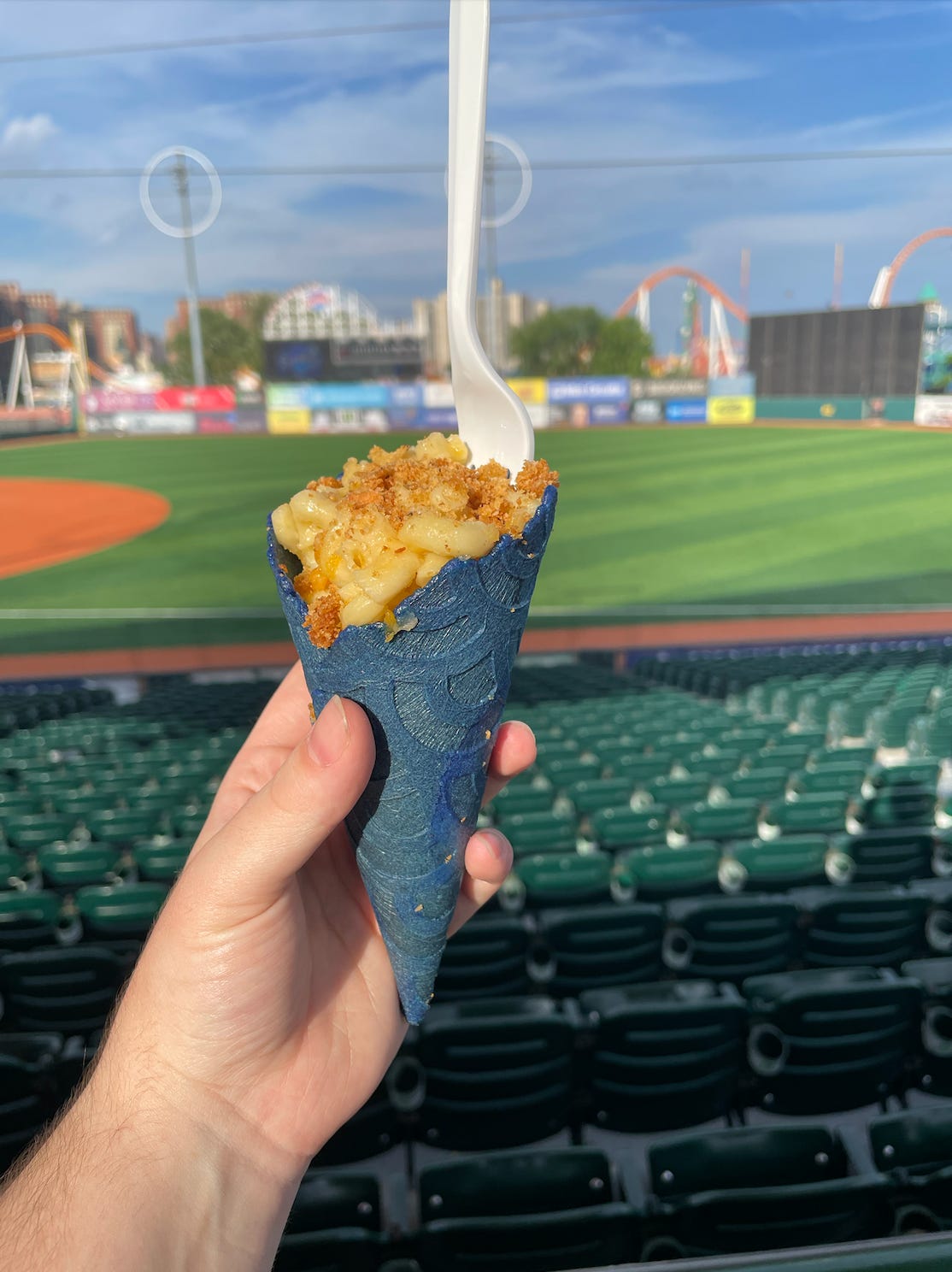 a hand holding a blue ice cream cone filled with mac and cheese, in front of a baseball field