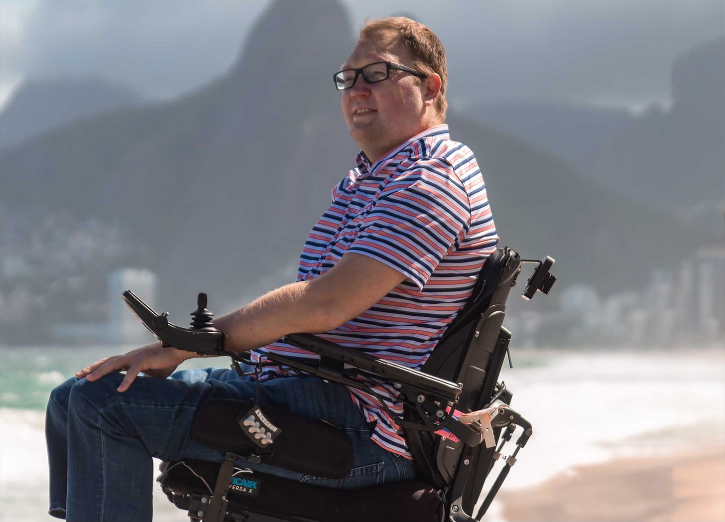 John seated in his wheelchair next to a beach with mountains in the background.