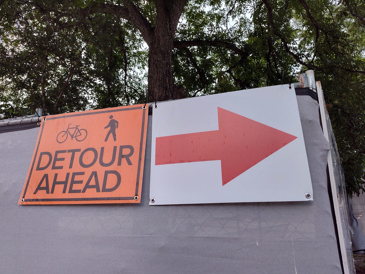 an orange sign with symbols of a bicycle and a pedestrian, with the words DETOUR AHEAD; a sign with a red arrow pointing to the right.