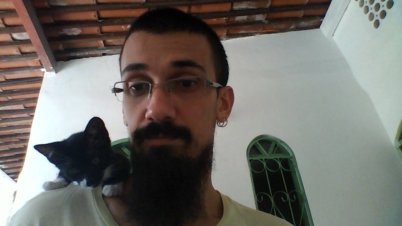 Selfie de mim com cabelo curto, óculos, barba e bigode pretos, e uma gata preta com focinho e patas brancas, ainda filhote, no meu ombro direito olhando para a câmera. Ao fundo, a parede e o telhado de uma casa.