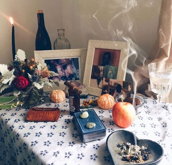 An altar decorated with flowers, framed photos of family, peeled oranges, an apple, cards, incense, water, candles, and rocks.