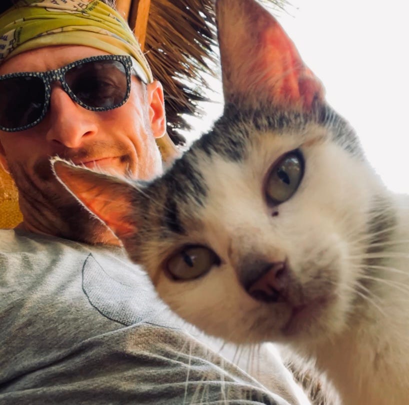 A man (the author) wearing sunglasses and a green bandana poses for a selfie with a stray cat, beneath a thatch-roofed building