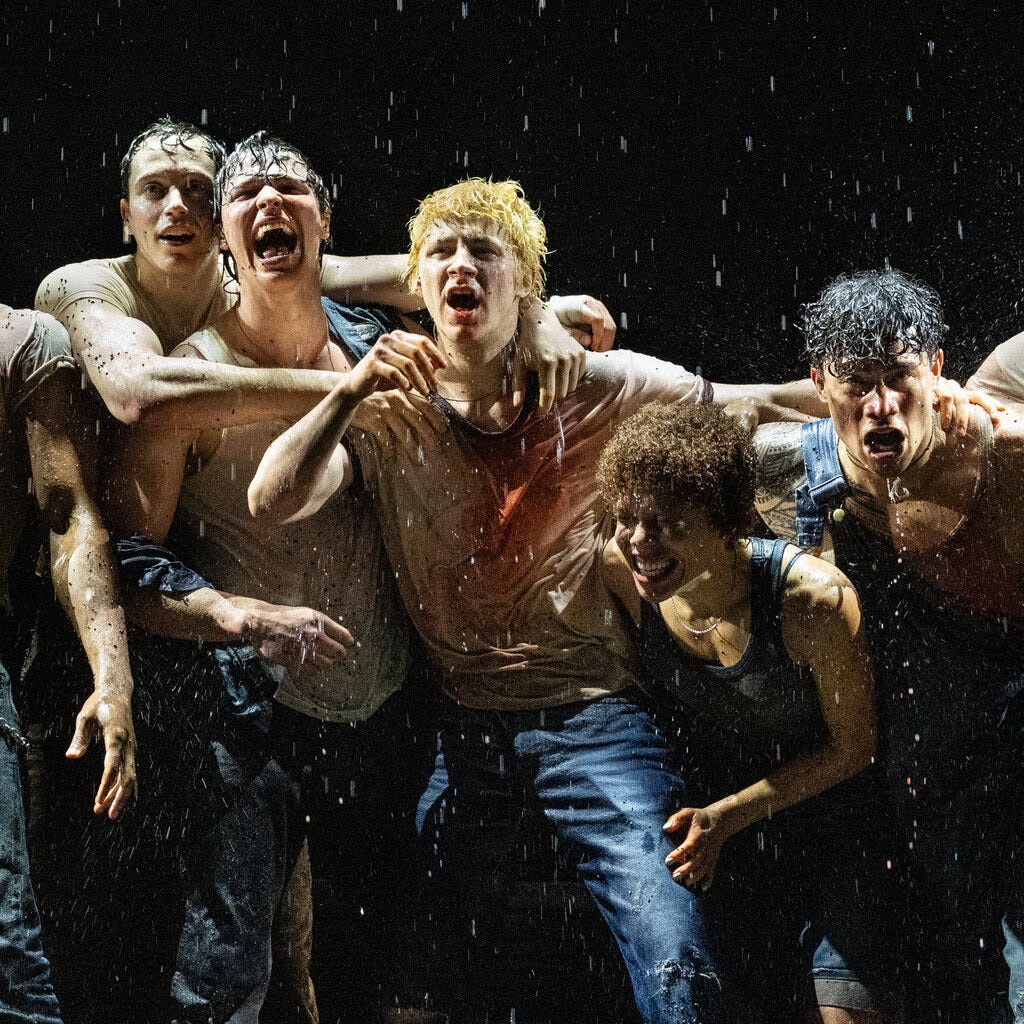 Several actors playing teenage boys are onstage in jeans and T-shirts with their arms around one another’s shoulders, shouting in a simulated downpour.
