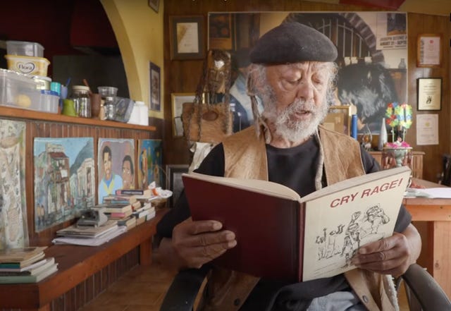 An elderly man in a beret with a great beard sits and reads from a book title 'Cry Rage'.