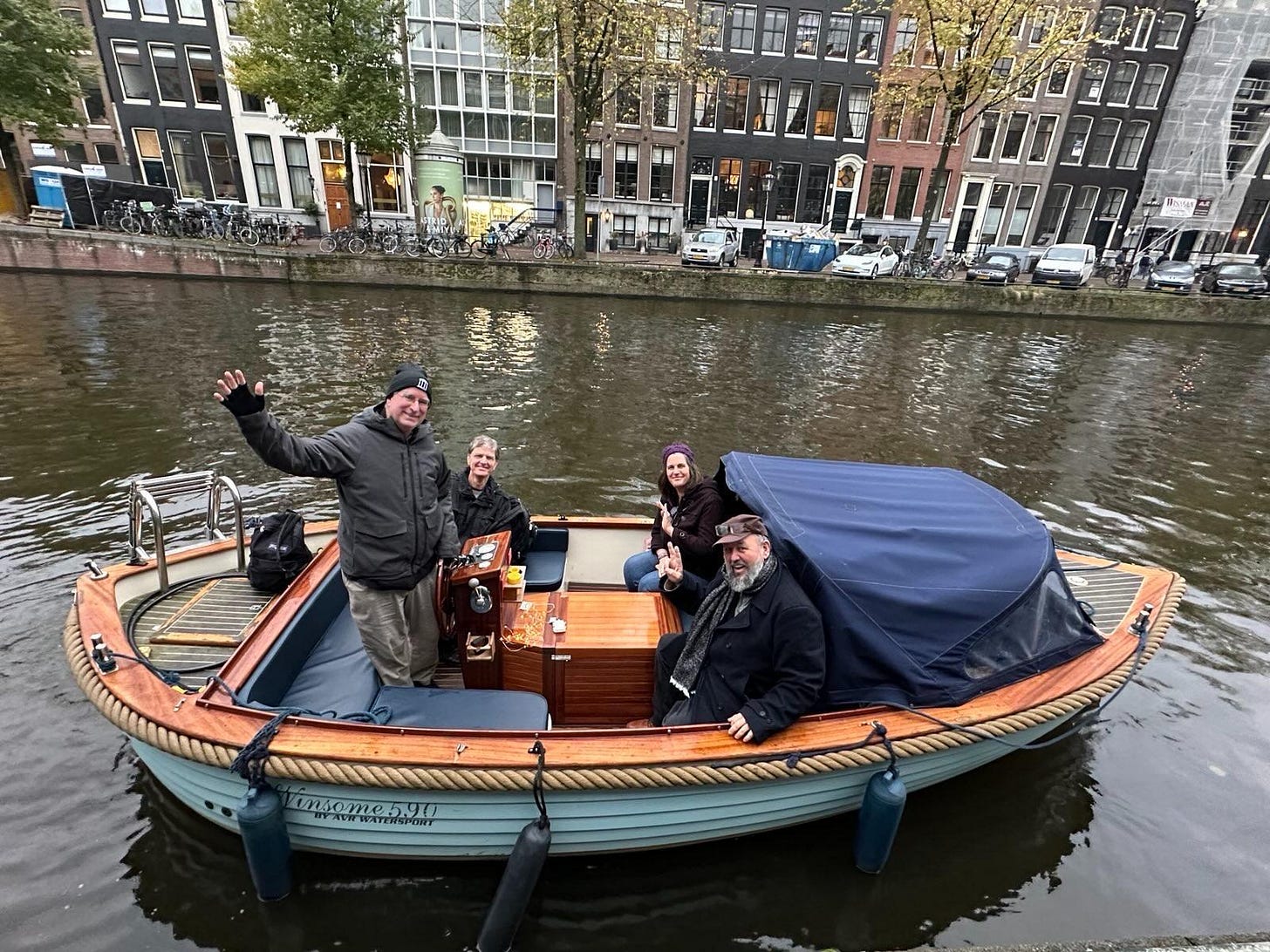  A boat in a canal in Amsterdam has four people all looking at the camera. A man stands at the wheel and waves. Three other people are seated. The canal takes up most of the shot, but there are typical Dutch canal houses in the background.