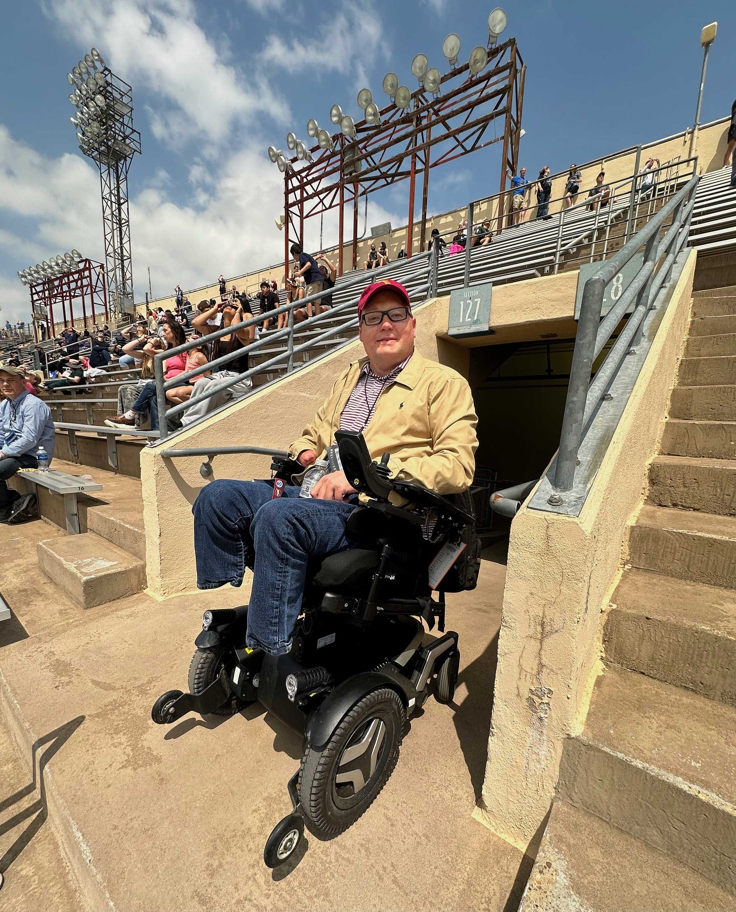 John seated in his wheelchair in a tunnel leading out onto the upper deck of seats at a stadium.