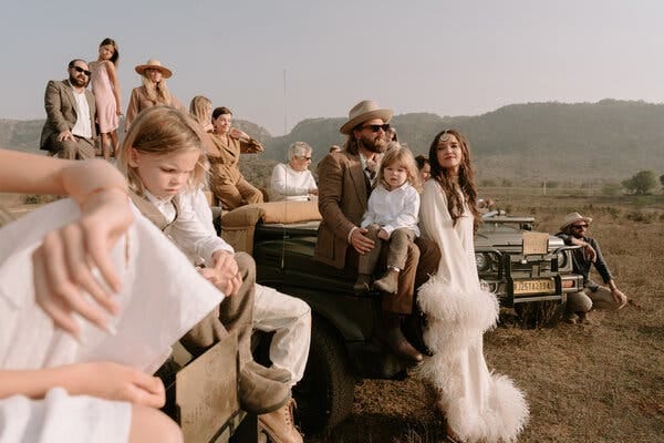 The couple surrounded by guests sitting, standing, squatting on and by jeeps, dressed in a color palette of tans, browns, whites and khakis, assemble on rugged terrain with hills in background for a safari wedding in India. The groom, left center, with a beard and sunglasses, wearing a taupe suit and a tan hat, sits on the hood of one jeep with a young child in his lap, wearing a white shirt and taupe khaki pants. The bride, standing next to them, with long brown hair, and wearing a headpiece down the center of her forehead, leans on the jeep, wearing a long white dress trimmed with puffs of white feathers.    