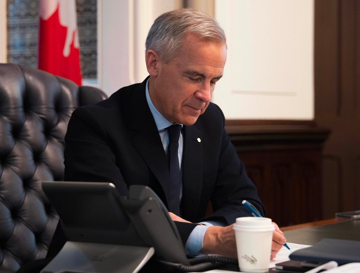 Prime Minister Mark Carney, wearing a suit, is seated at a desk with Canadian flags displayed behind him. He is looking down and writing notes on a paper.