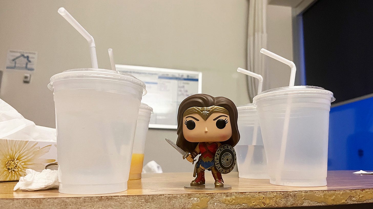 Wonder Woman funko pop toy figure standing amid a bunch of empty plastic cups with bent straws on a patient hospital tray table.