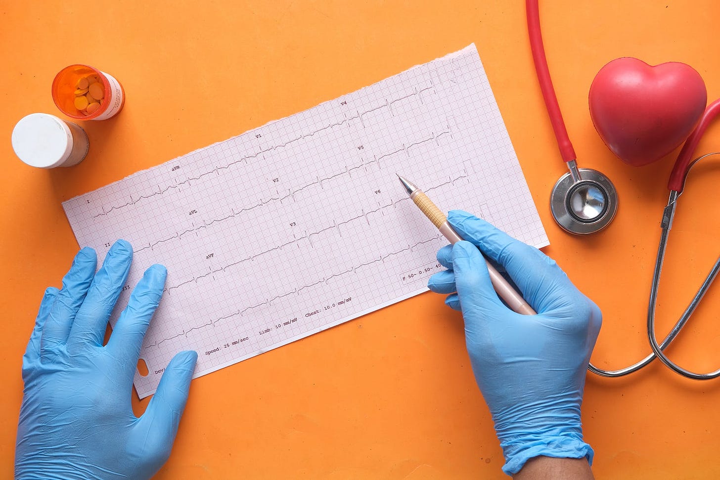a top-down view of pair of hands wearing blue medical gloves writing on a medical document on an orange background. Two pill boxes sit on the top left of the image and there is a stephoscope on the right sitting next to a red heart-shaped object.