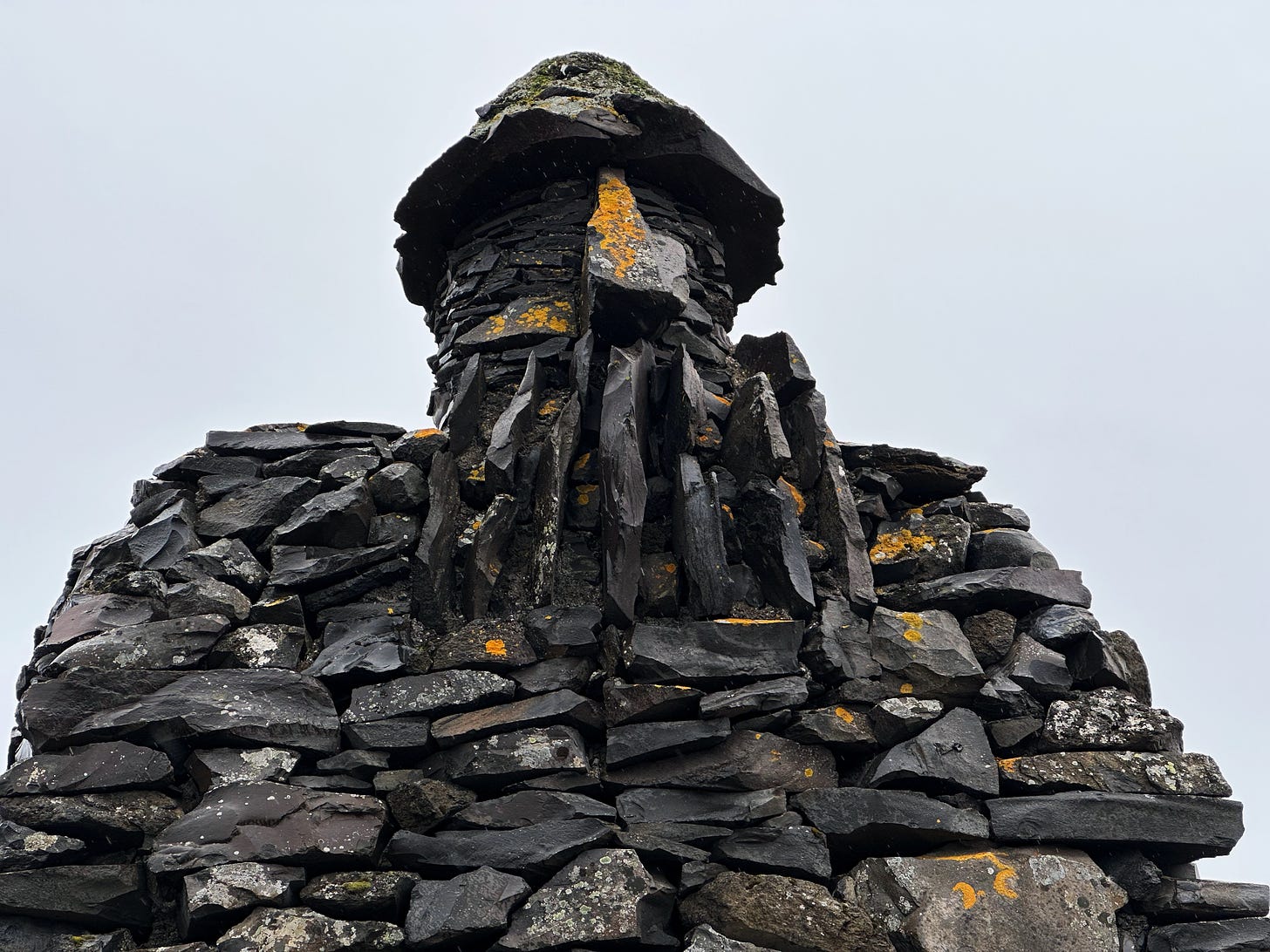 A close up view of the top half of Bárður Snæfellsás a large stone sculpture of a stone troll called the protector of the Snaefellsnes Peninsula. It is constructed of small pieces of dark rock covered in lichen and moss and shown against a blue gray sky.