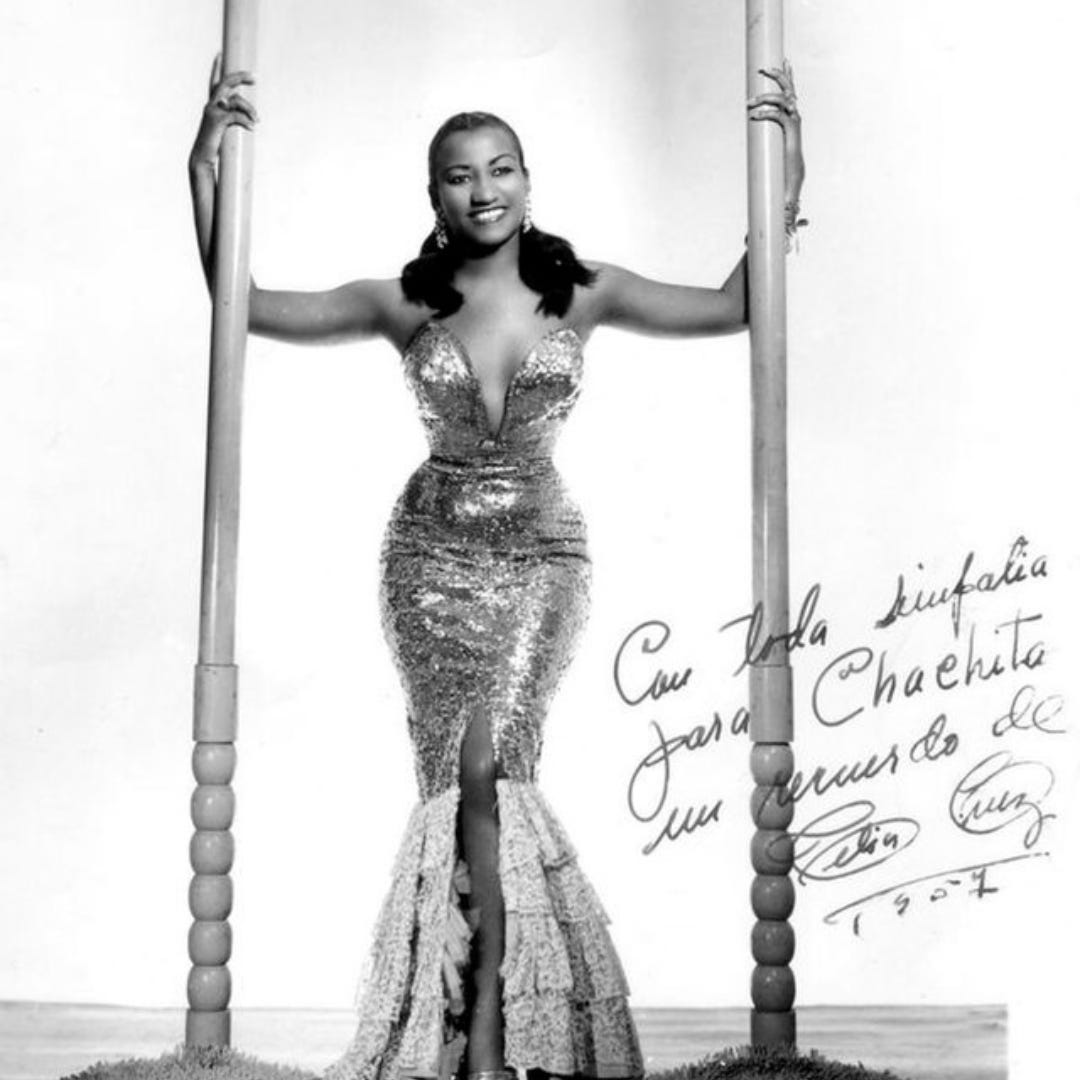 A black and white image of Celia Cruz in her early years. She looks glamorous in a sequin dress with a plunge neckline that shows off her hourglass figure. She smile a big smile at the camera. 