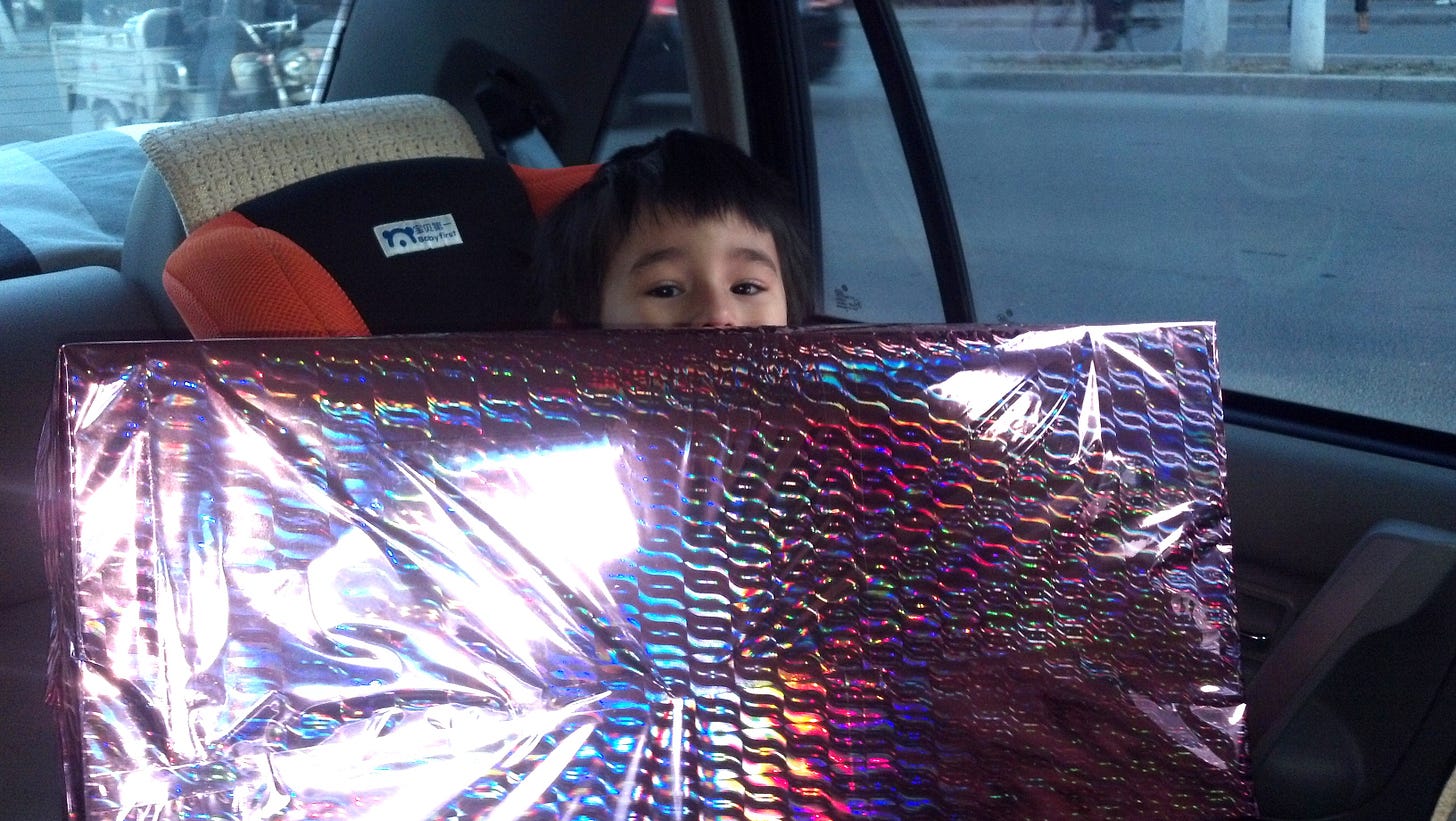 A small child in a car safety seat peering over a large box wrapped in irridescent paper.