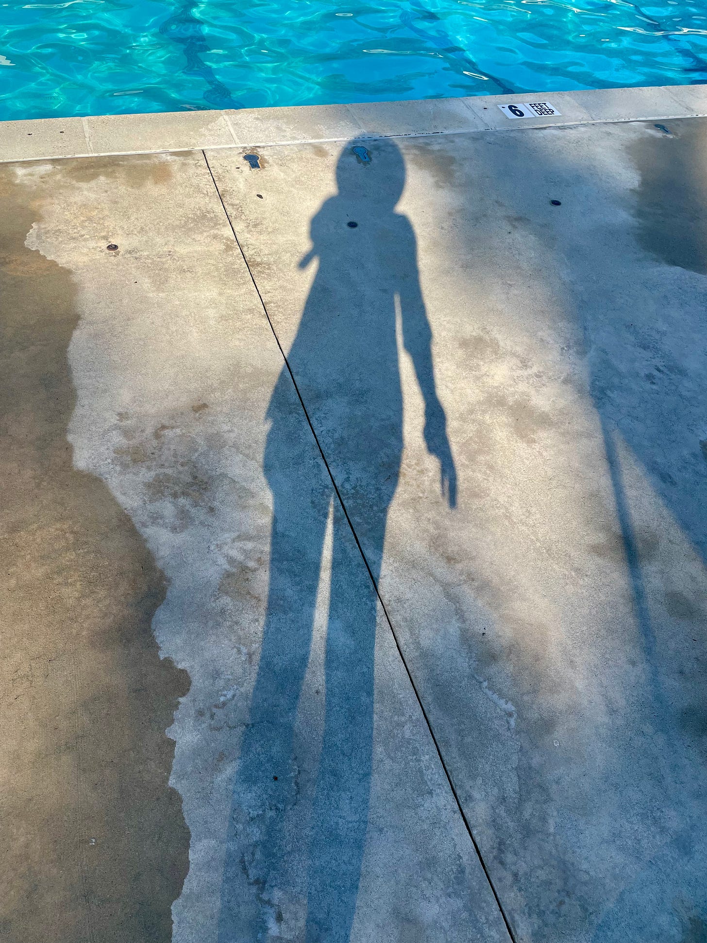 A photo of my full-body shadow on the concrete of a pool. Water marks are to my left. The ripple of the pool is in the upper inch of the frame.