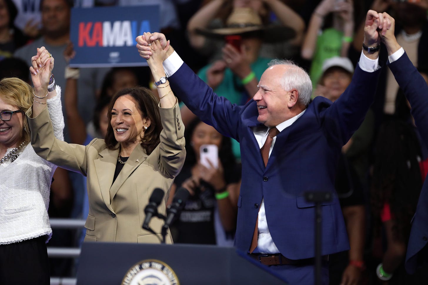 Kamala Harris and Tim Walz raising their hands in the air together with supporters holding signs behind them