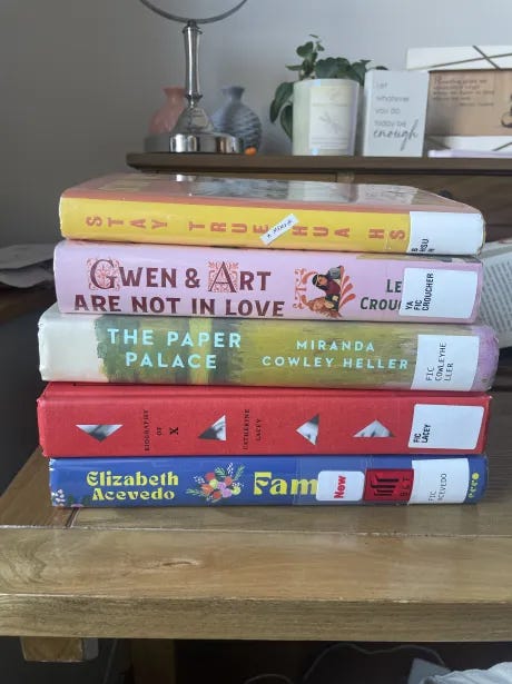 Stack of hardcover books on a wooden desk, from top to bottom: Stay True by Hua Hsu, Gwen & Art Are Not in Love by Lex Croucher, The Paper Palace by Miranda Cowley Heller, Biography of X by Catherine Lacey, and Family Lore by Elizabeth Acevedo.