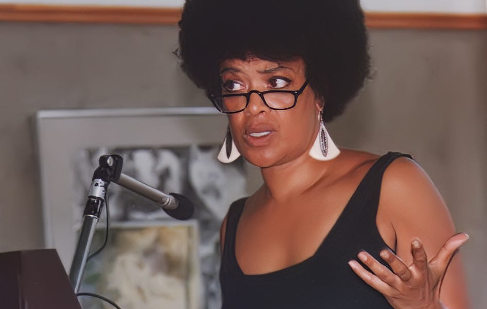 toni cade bambara speaks in front of a microphone, black tank top, black afro, black rectangular glasses on the bridge of her nose, smooth black skin and white triangle earrings dangle as she gesticulates with her left hand and arches her right brow.