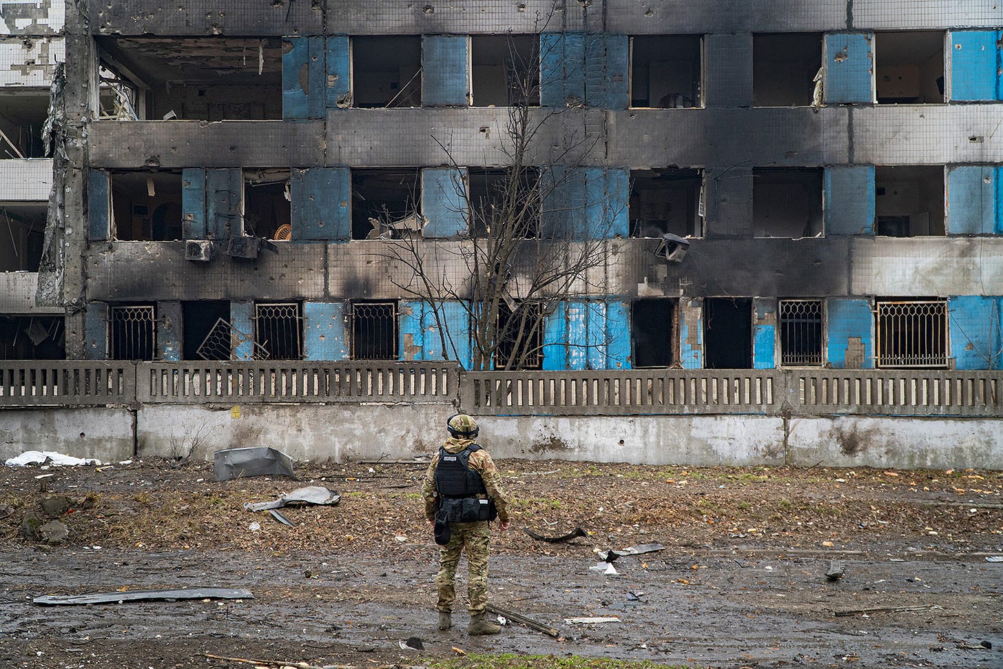 A Ukrainian police officer in full uniform and protective vest and helmet stands in front of a maternity hospital damaged by Russian shelling. The windows are blown out and burned and the debris litters the ground.