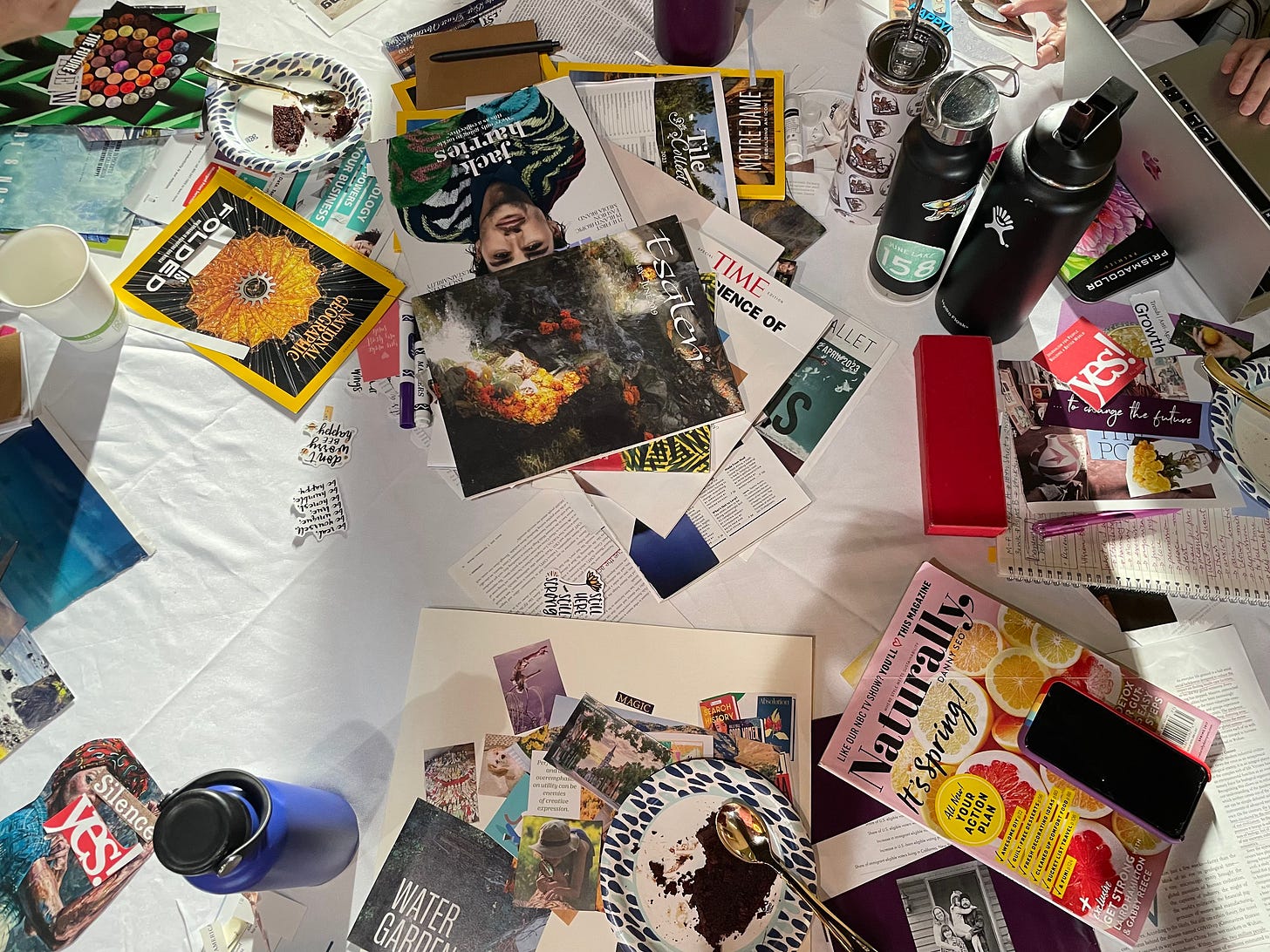 A table with magazines, images cut out from the pages, and poster boards scattered throughout. Water bottles, phones, and plates with cake are also seen. 