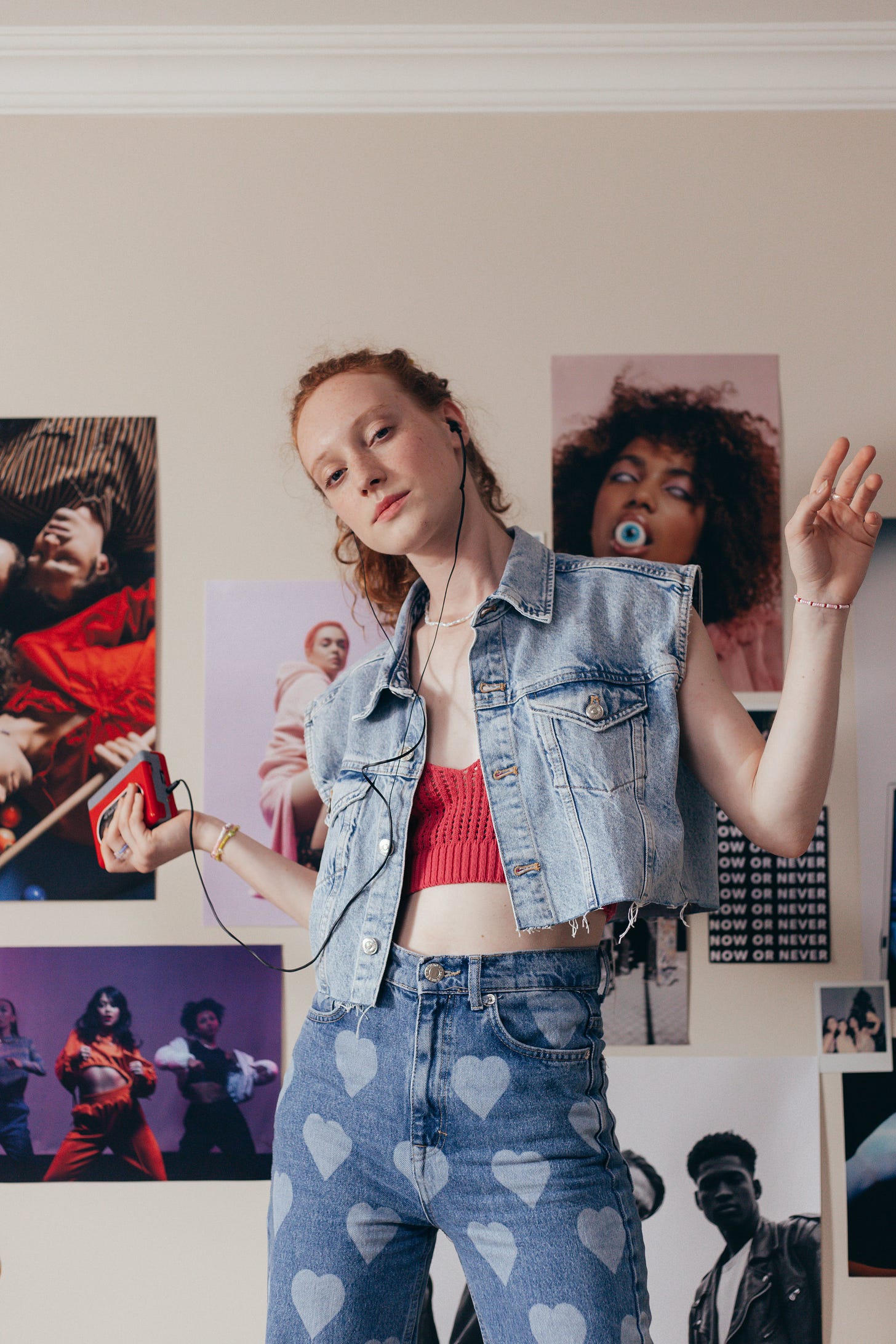 A young redheaded white woman in a jean vest and jeans with hearts on them listening to a walkman in front of a bunch of band posters.