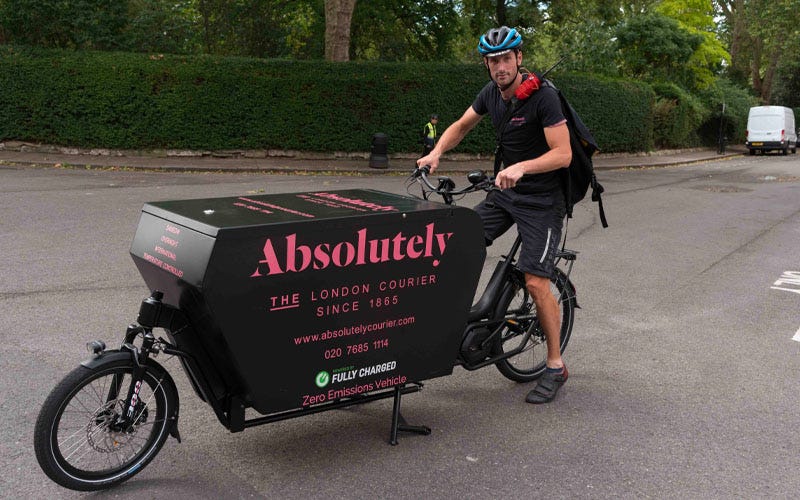 A cycle courier rides a modern cargobike along a street in London. The bike is black with the name of the courier company on the side