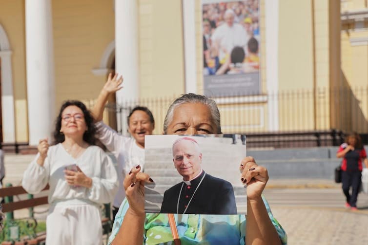 A woman holds a photograph of a man wearing a black clerical coat, while two others stand beside her and wave. A woman holds a photograph of a man wearing a black clerical coat, while two others stand beside her and wave.