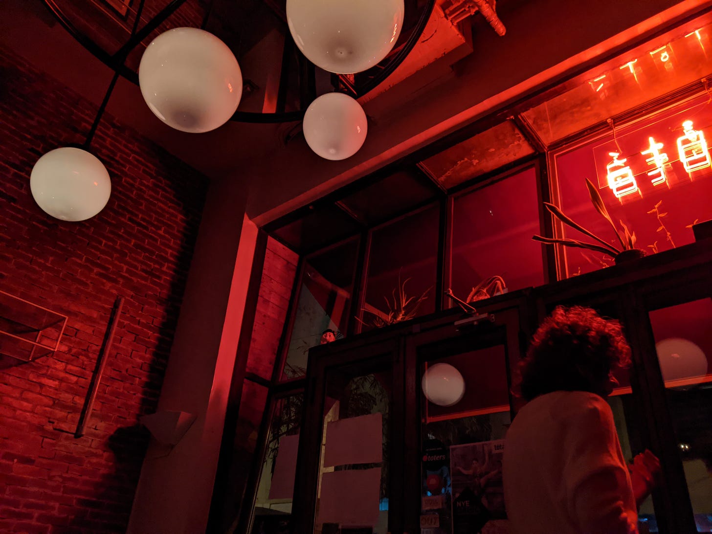 View from inside a bar. White globe lights hang from the ceiling, and fluorescent lights in the shape of Chinese lettering adorn the outer window.