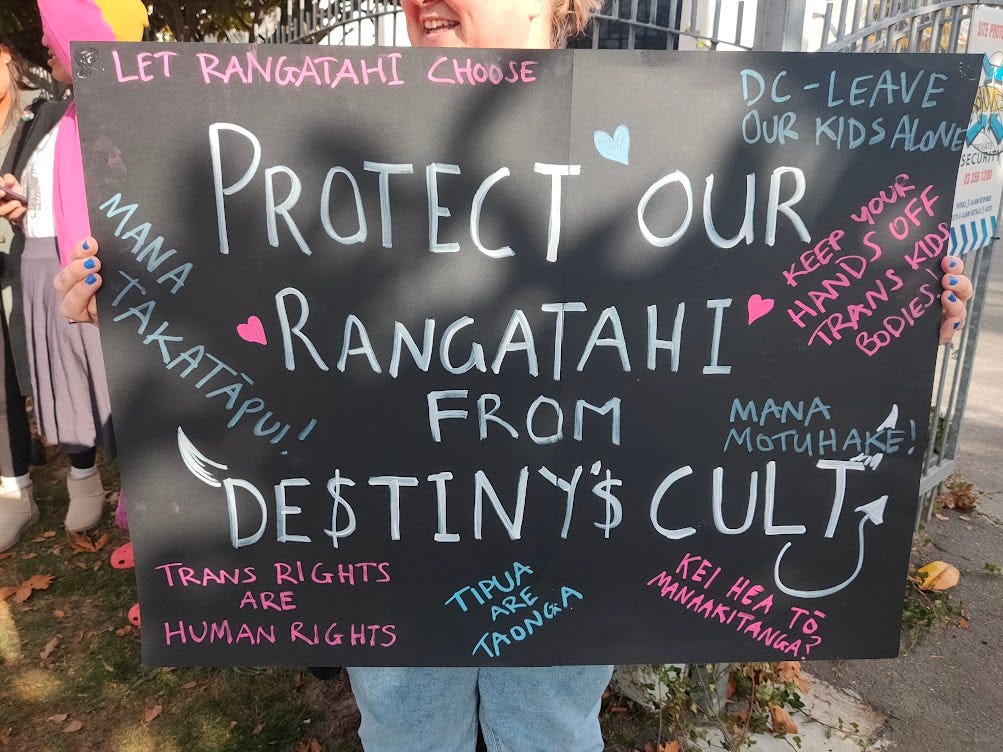 a woman holds up a sign reading 'protecy our rangatahi from Destiny's cult'