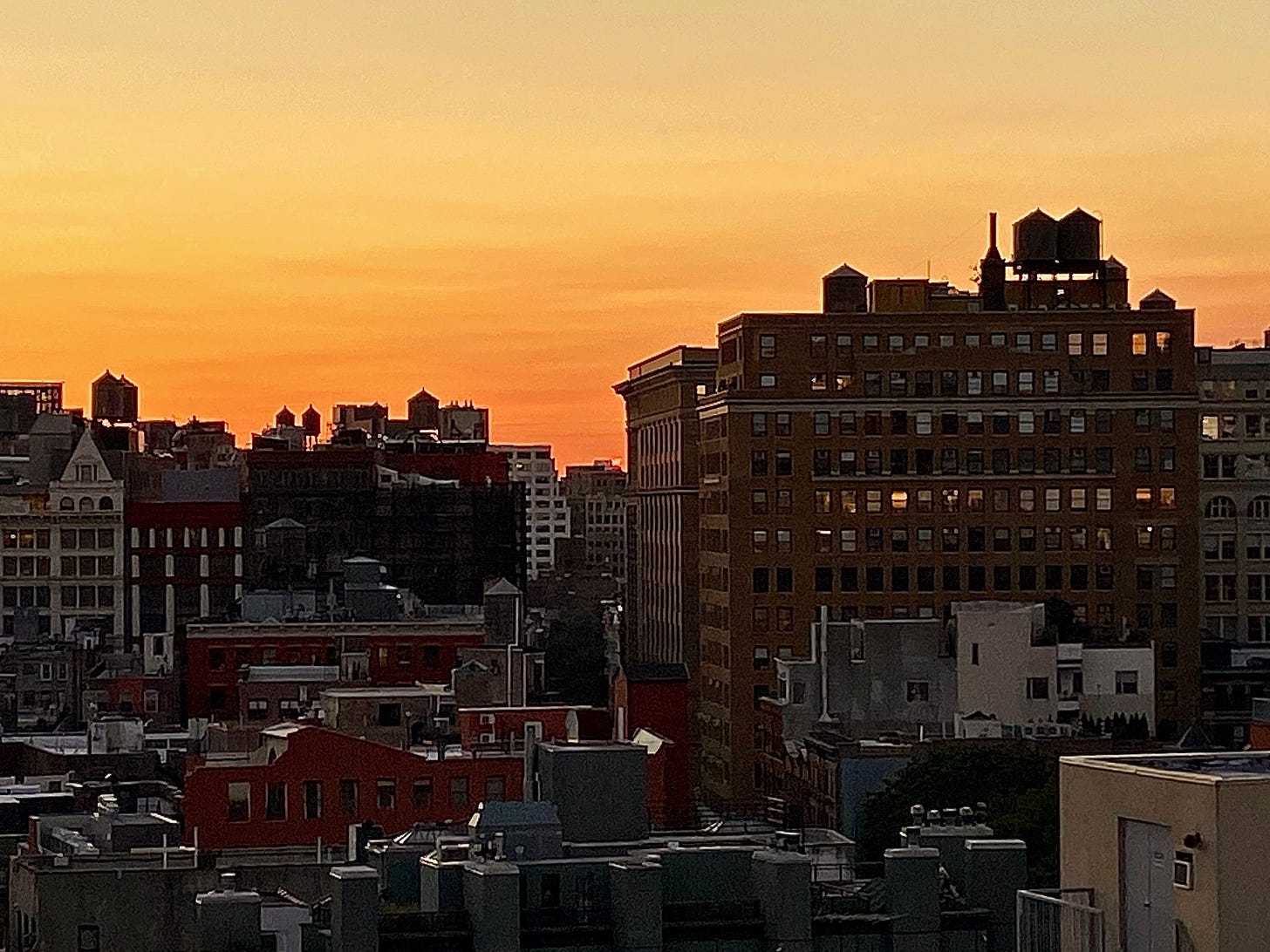 The sun setting behind the skyline of the lower east side. Multiple water towers of different sizes and in different formations are silhouetted against the orange of the sky.