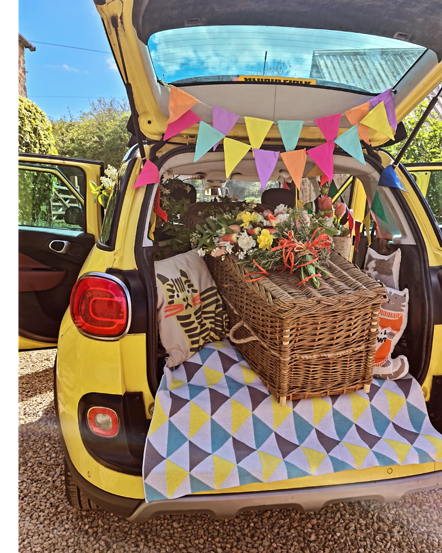 The open boot of a yellow car decorated with colourful bunting, cushions and blankets, a willow coffin topped with fresh flowers