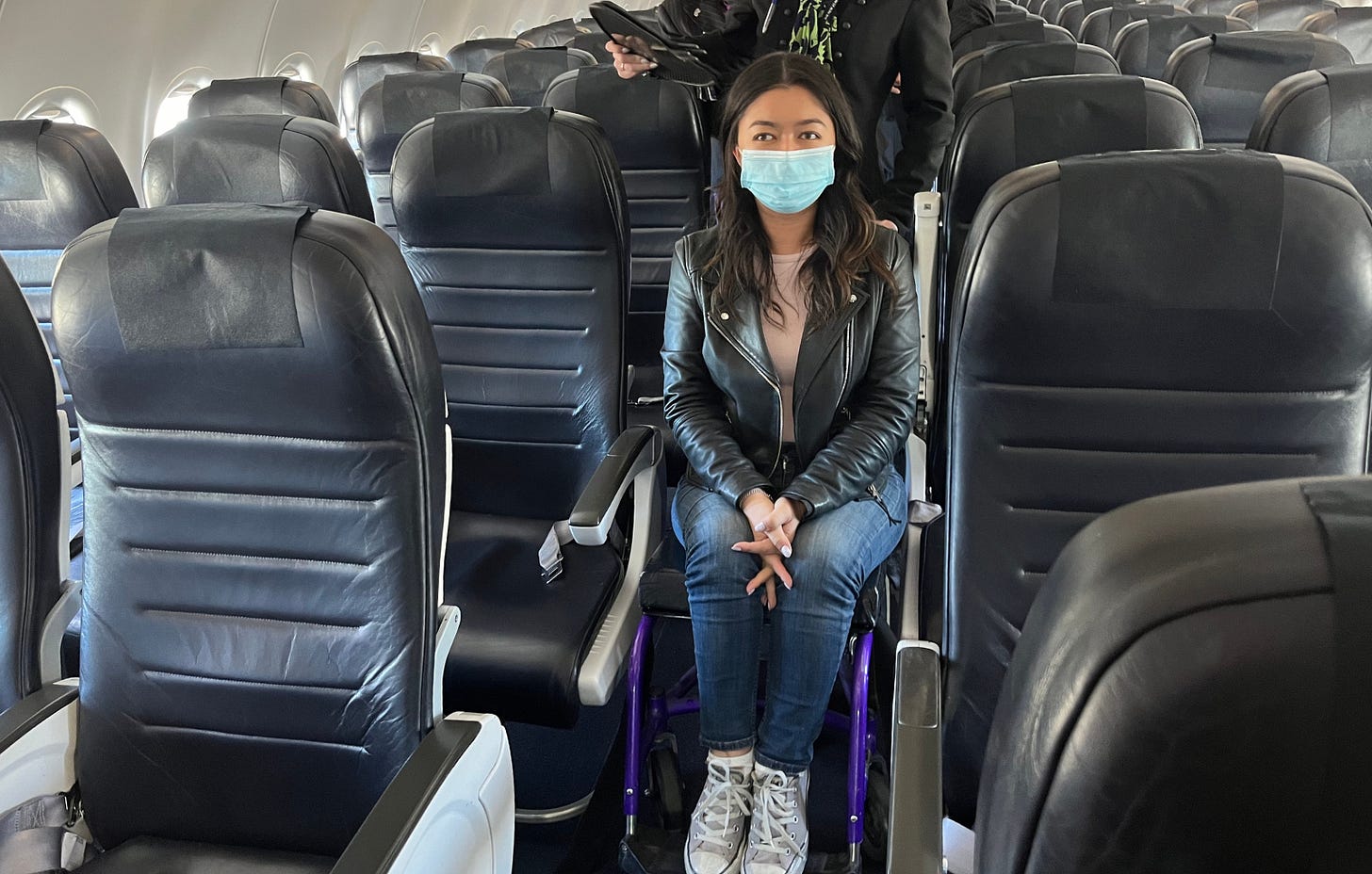 Olivia, wearing a black jacket and face mask, sits in an aisle chair in the middle of an aeroplane. she is surrounded on either side by rows of empty seats.