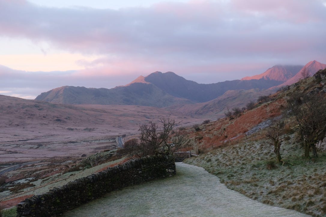 A pink and purple sunrise casts a soft glow over mountains in Eryri National Park, Wales. A driveway is visible in the foreground.