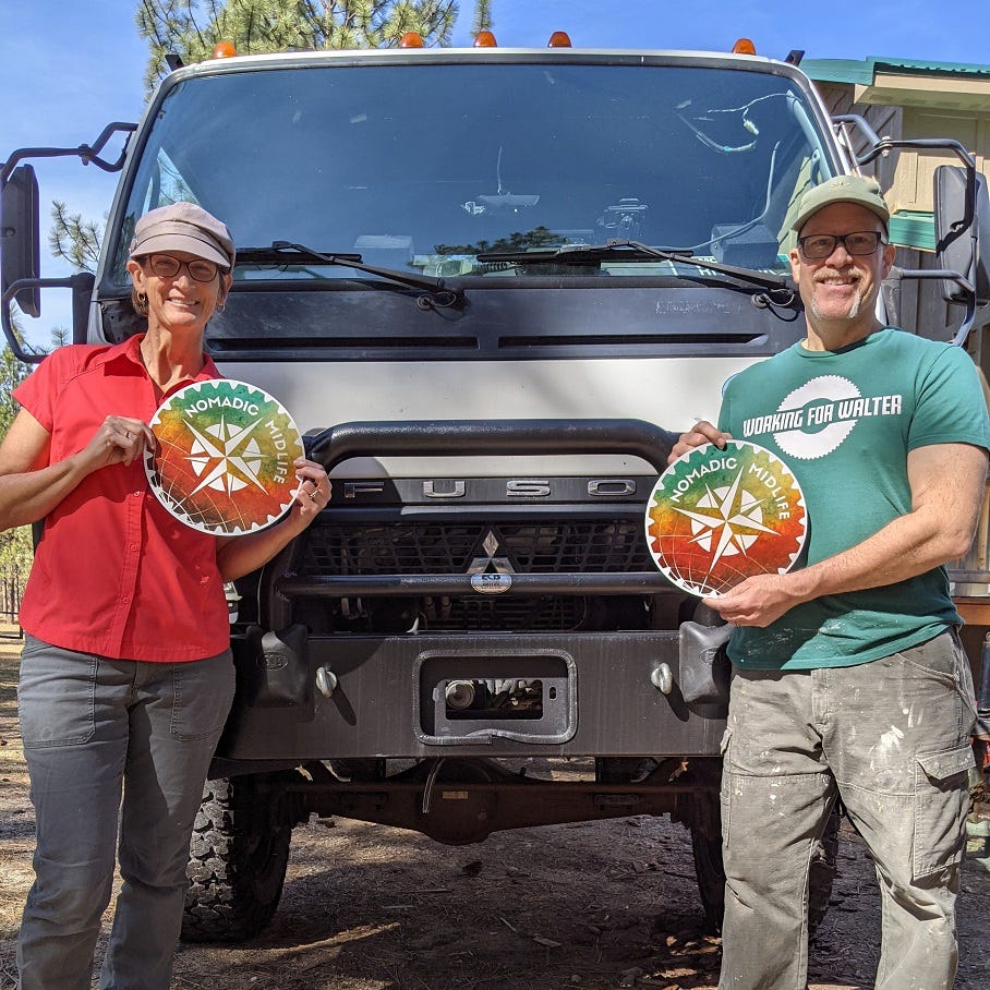 Andy and Sherry are each holding a 12 inch round magnet with their Nomadic MidLife logo, standing in front of their Mitsubishi Fuso truck