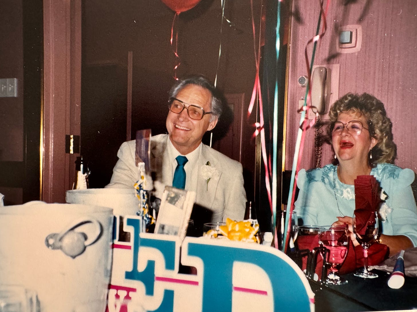 A 50-something-year-old white man and his wife sit smiling amongst party favors celebrating the man's retirement.