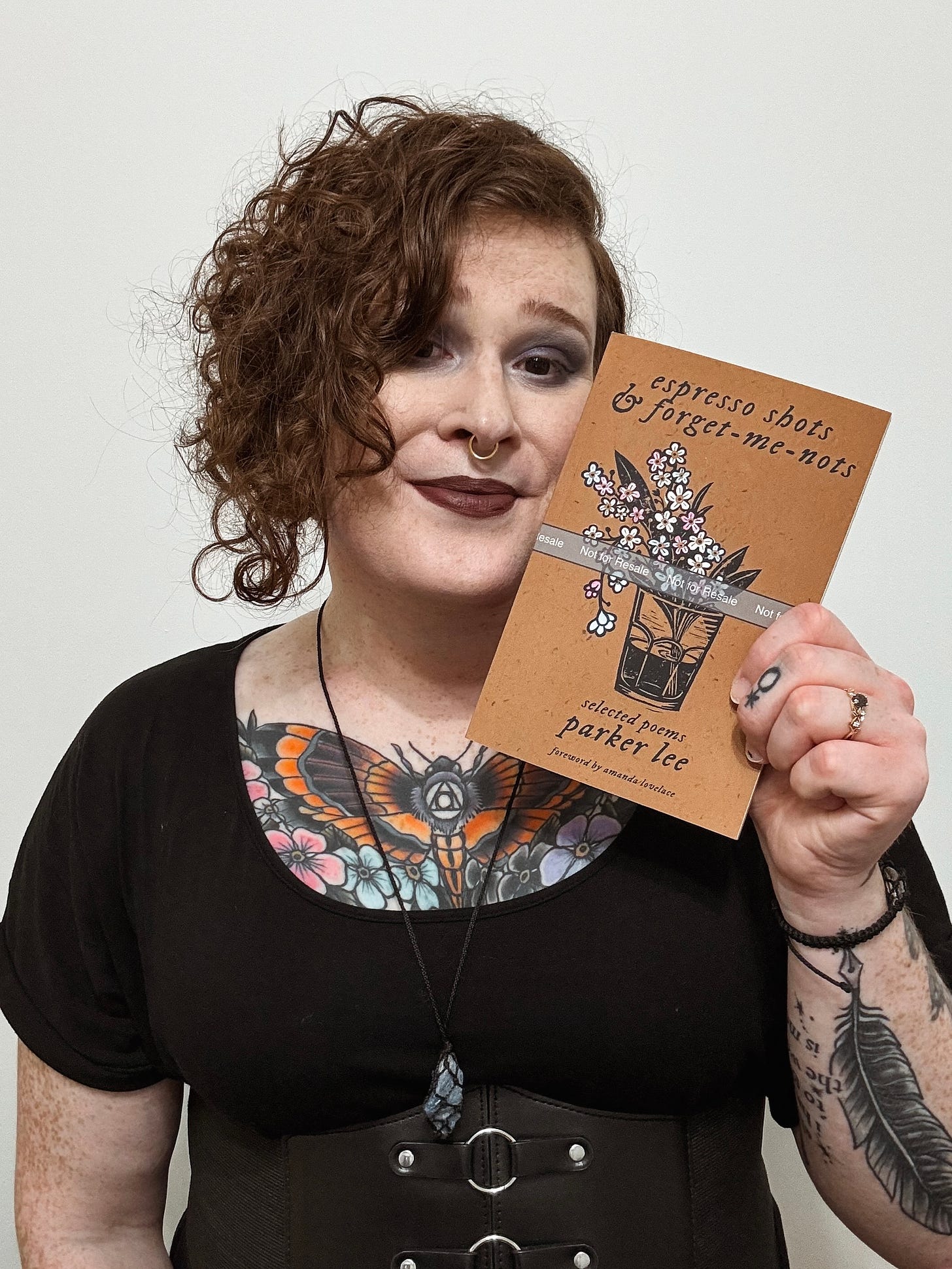 a photo of Parker from the waist up holding a copy of “espresso shots & forget-me-nots.” they are wearing a black scoop neck dress with a black corset belt at her waist, and a black cord necklace with a piece of sodalite hanging from it.