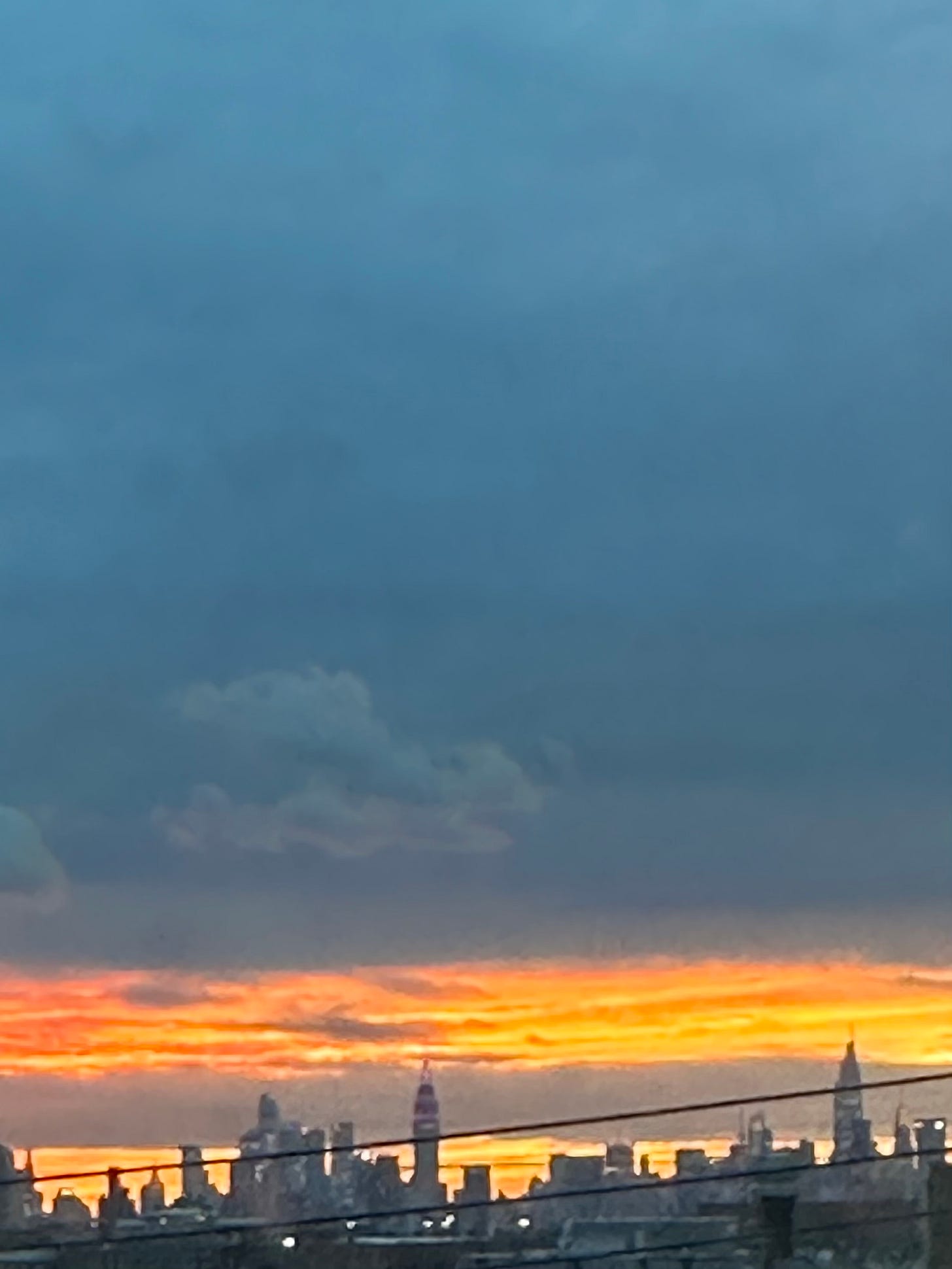 New York City at dusk, a mostly blue and gray sky with a bright orange and yellow sunset near the bottom of the frame, and a portion of the skyline