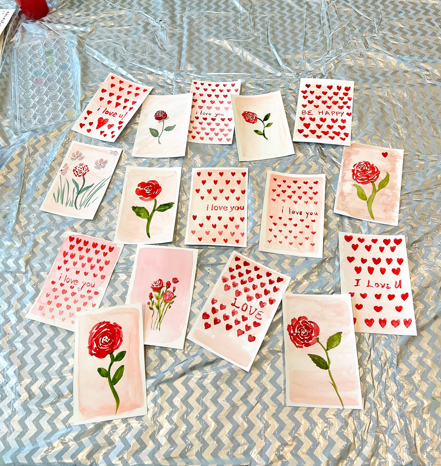 display of multiple watercolor paintings to be used as Valentine's Day cards, in two designs, one of a single stem rose in a loose watercolor style and one with hearts and lettering