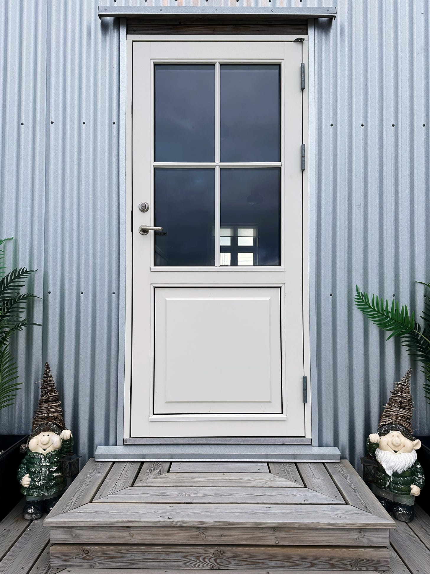 The front door of our rental home.  The door is white with large windows, the siding is silver metal and there are two ceramic gnomes in green coats welcoming us as we approach.