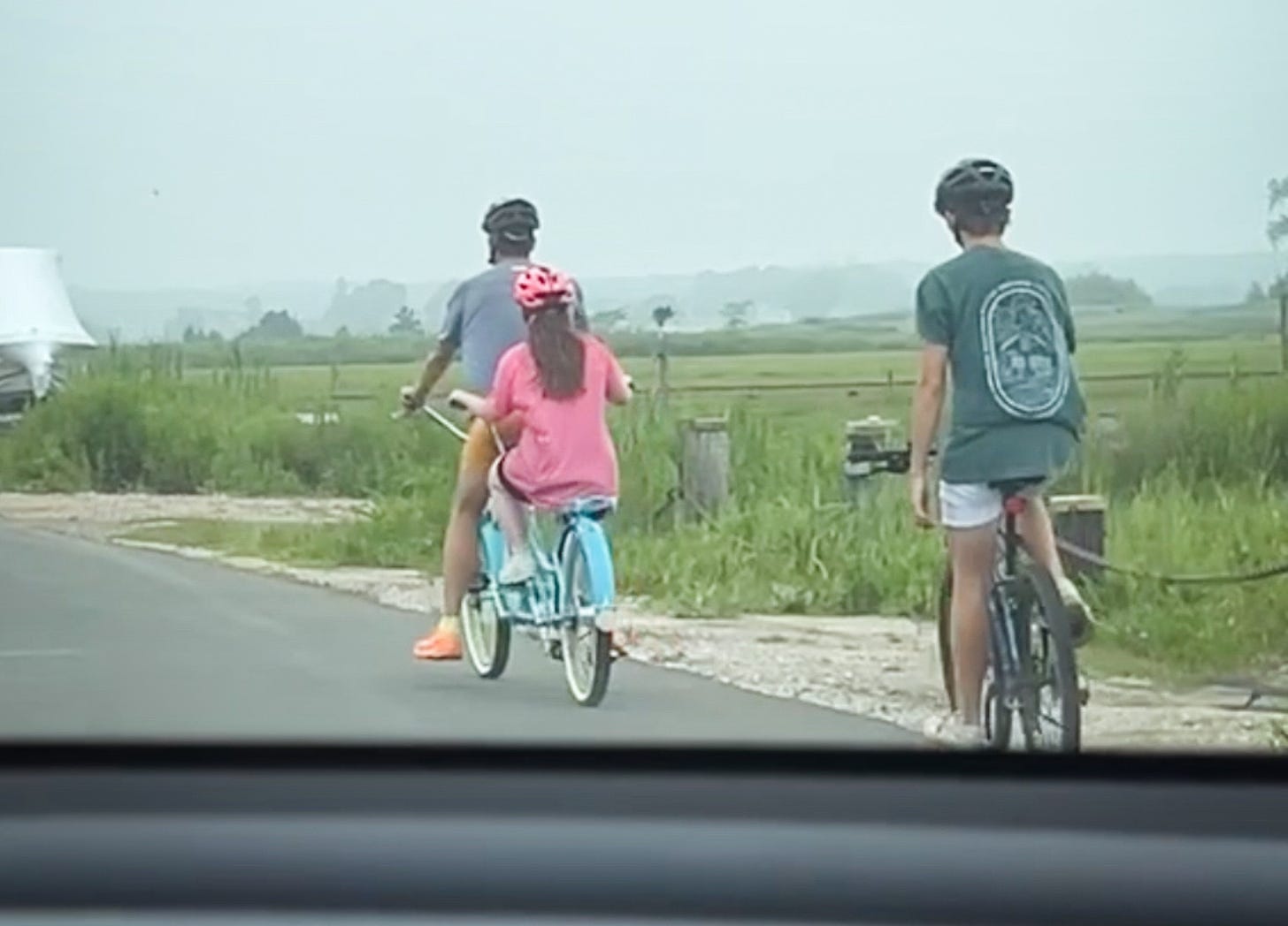 A blurry photo taken from a car of Penny pedaling on a tandem bike behind Peter. William is on another bike behind them.