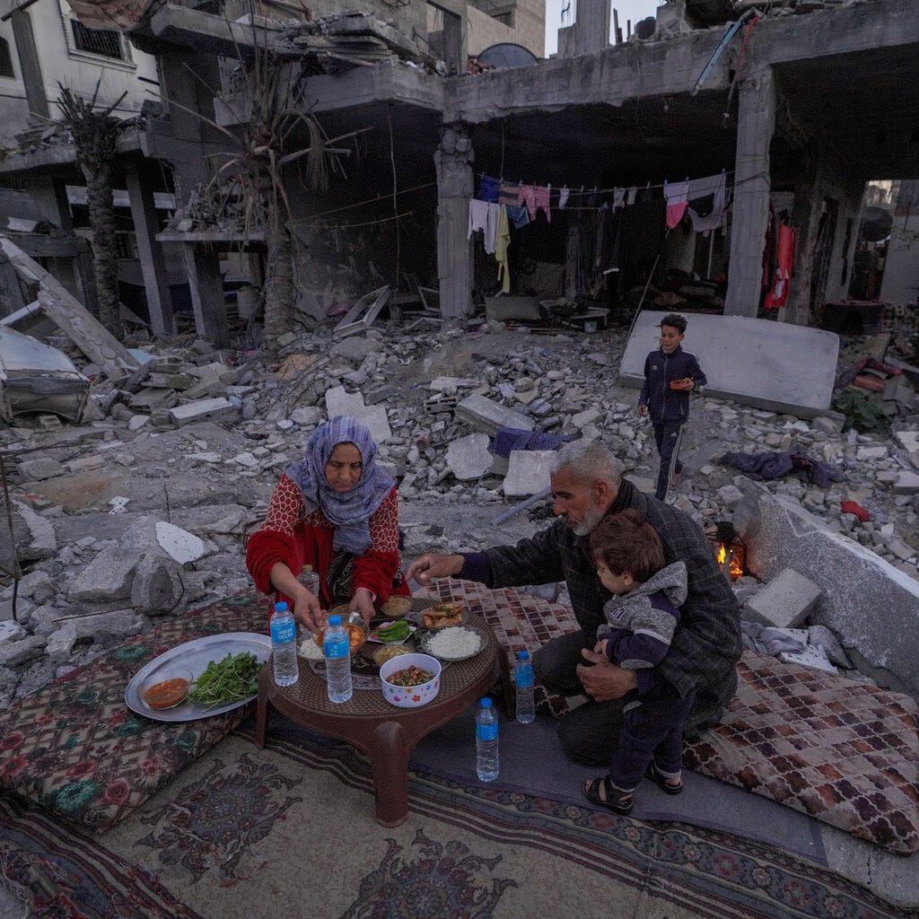 A family setting up a meal on a carpet over rubble, surrounded by destroyed buildings.