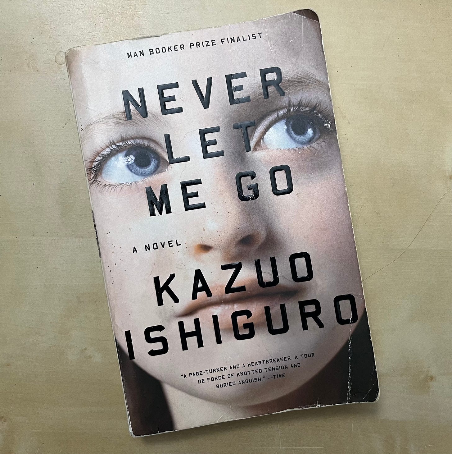 Photo of my paperback copy of Never Let Me Go by Kazuo Ishiguro, which has a close-up picture of a female-presenting face on the cover, a white young adult with big blue eyes looking off to the side